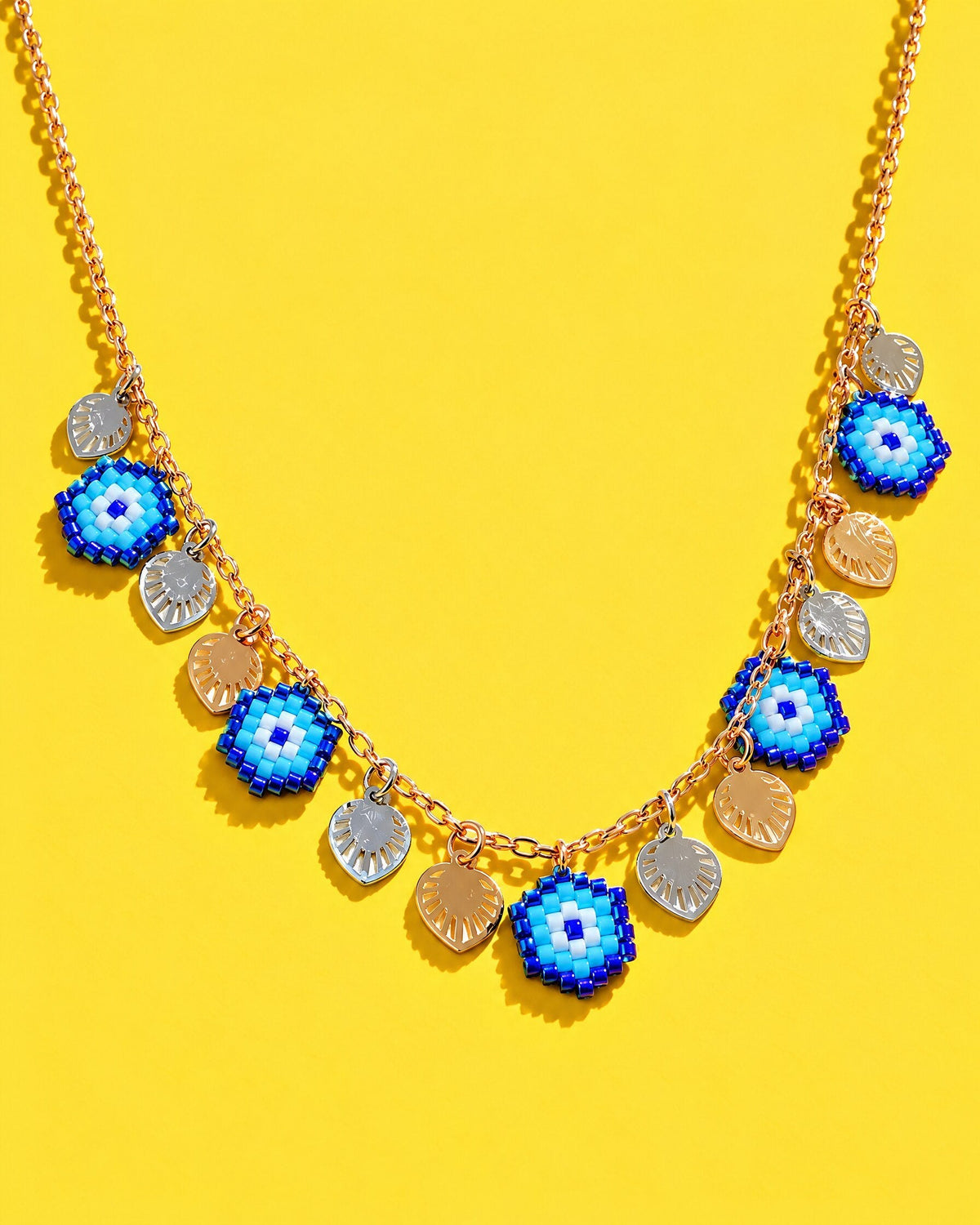 A beaded evil eye charm necklace with cobalt, turquoise and white eyes and gold and silver leaf charms on a yellow background